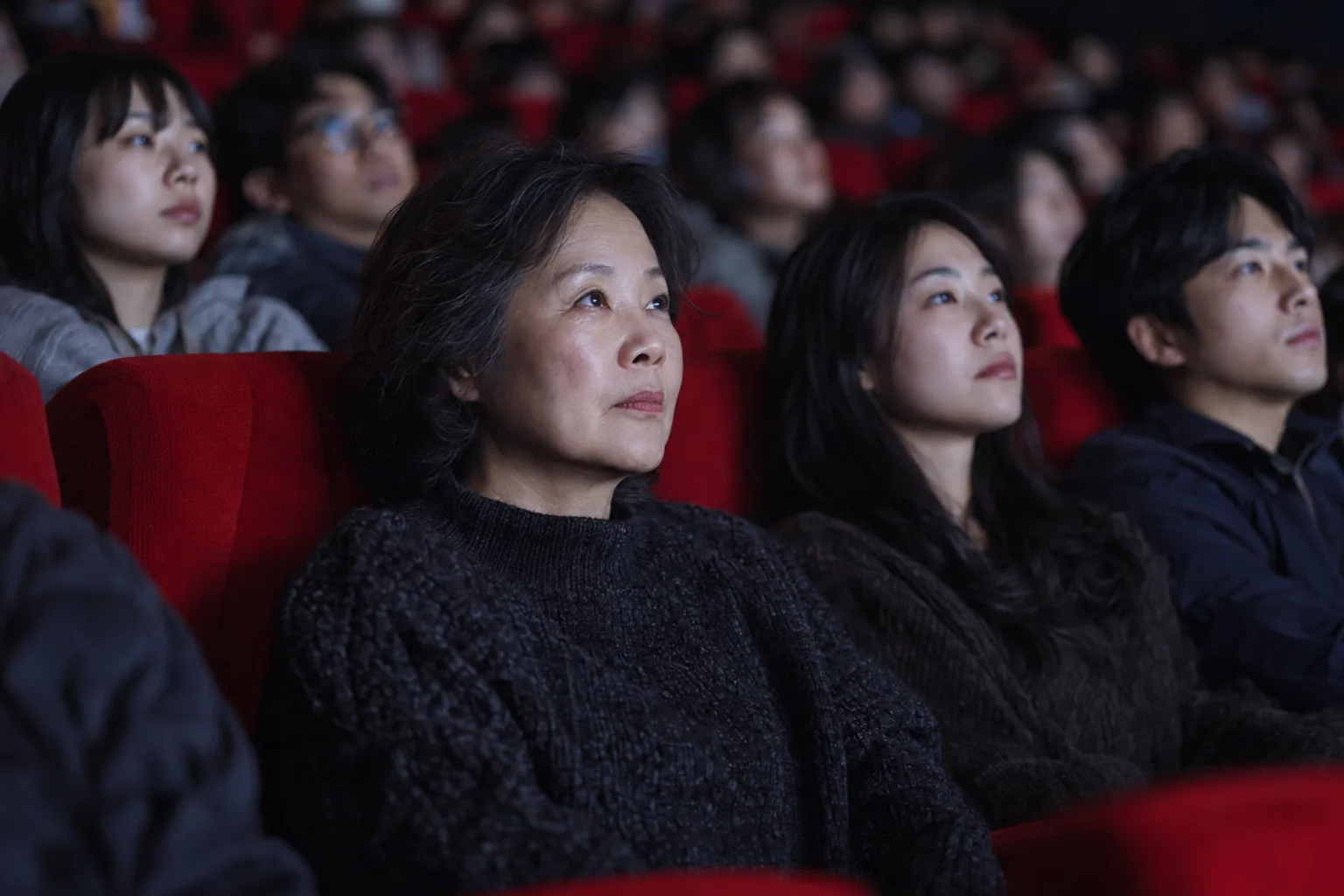 赤い座席が埋まった映画館で、暗い色のセーターを着た年配の女性がスクリーンを見つめる。