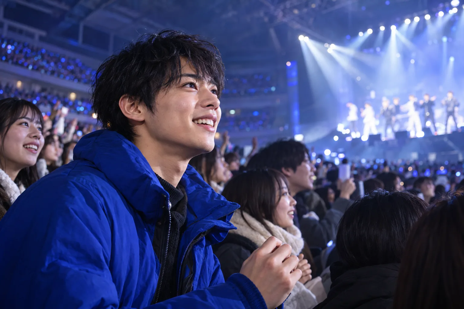 A young man in a blue jacket smiles toward a brightly lit stage amid an indoor arena crowd.
