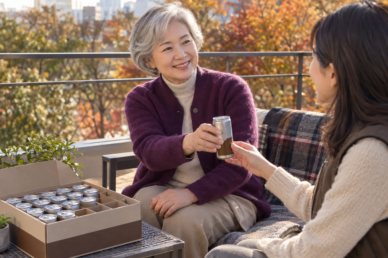 A silver-haired woman in a purple cardigan hands another woman a small can beside an open box of cans on a sunny balcony.