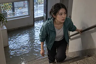 雨水が床の排水口の周りに広がる半地下の部屋から、女性が階段を上る。