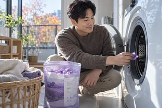 A man places a purple detergent capsule into an open washer beside a pouch of capsules and a basket of towels.