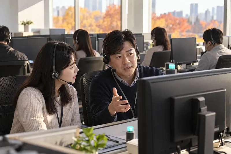 Two headset-wearing support staff confer over a monitor, with a teal light glowing at their desk and other agents behind them.